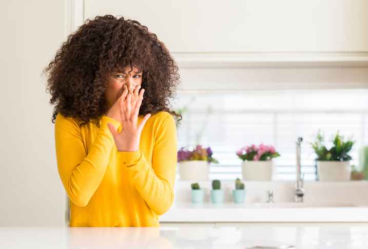 Adult woman wearing yellow sweater at kitchen smelling something stinky and disgusting, intolerable smell, holding breath with fingers on nose. Bad smells concept.