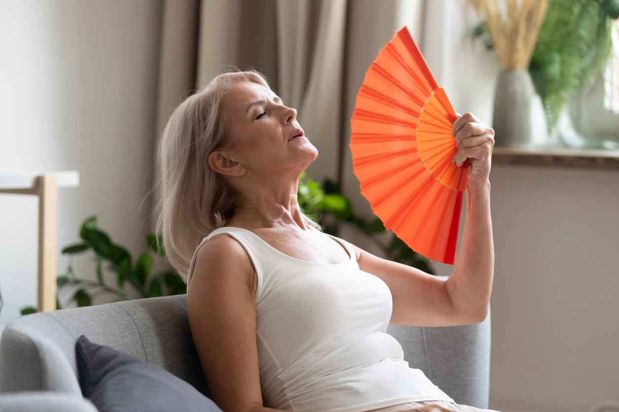 Stressed annoyed old senior woman using waving fan suffer from overheating, summer heat health hormone problem, no air conditioner at home sit on sofa feel exhaustion dehydration heatstroke concept